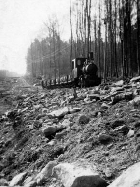 Auf der schwarzweiß Aufnahme ist eine Dampflokomotive mit Muldenkipper zu sehen.